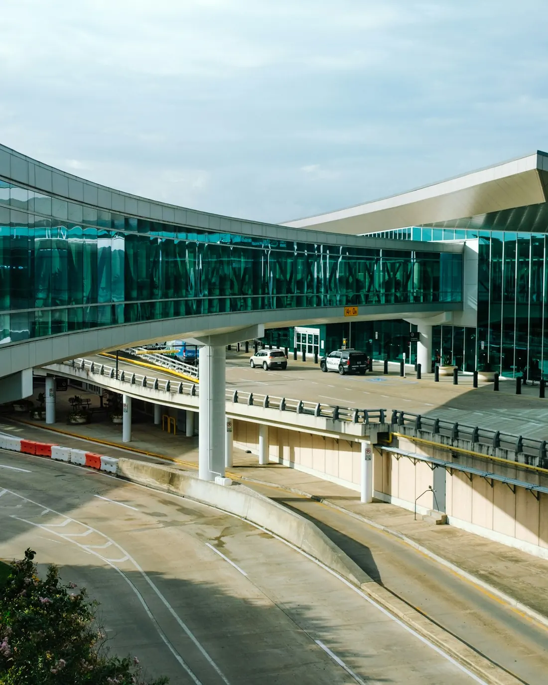 An airport terminal seen from outside, with a curved glass walkway running above the pickup roadway.