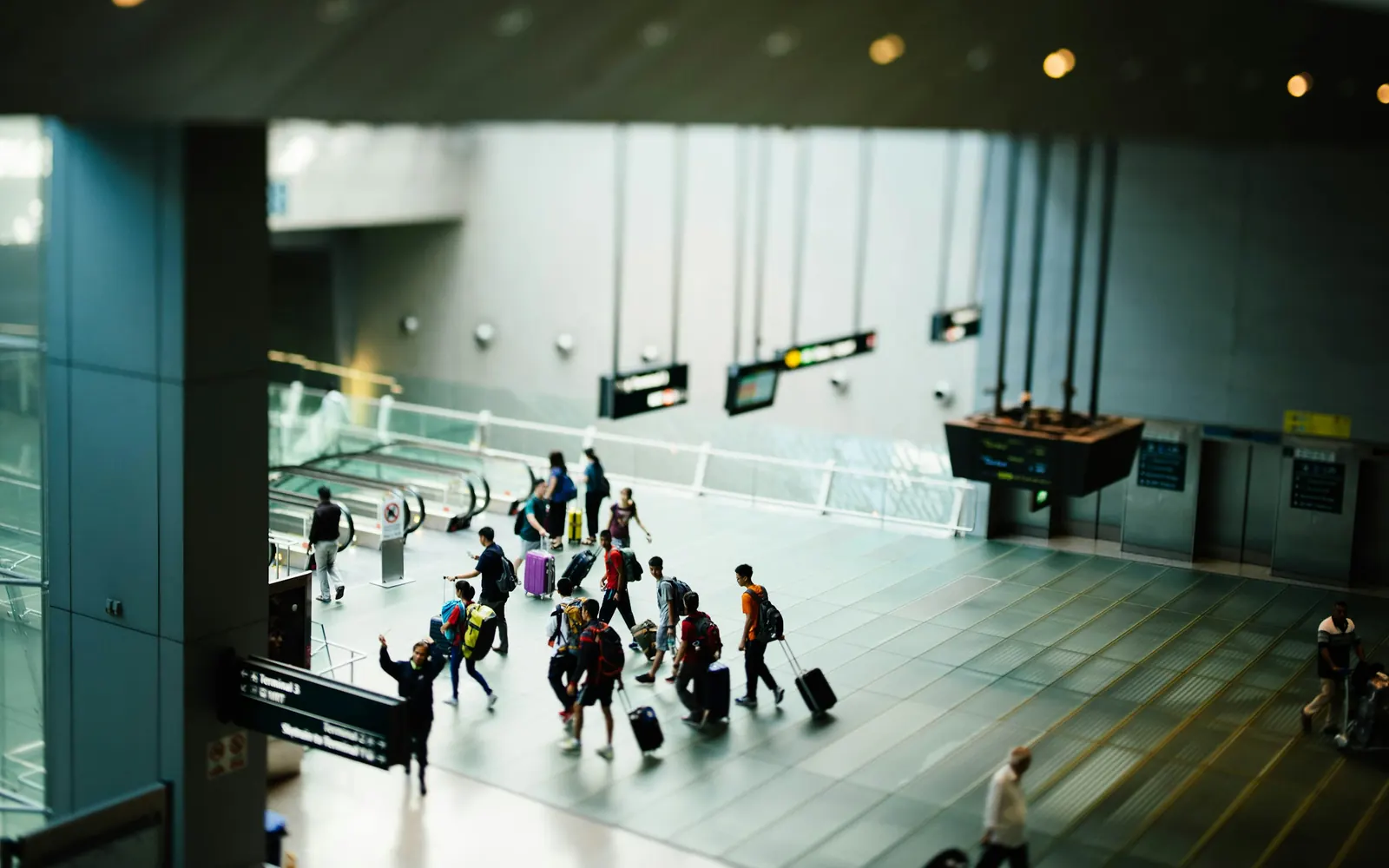 Travellers wheeling suitcases across a wide airport concourse, seen from a floor above.