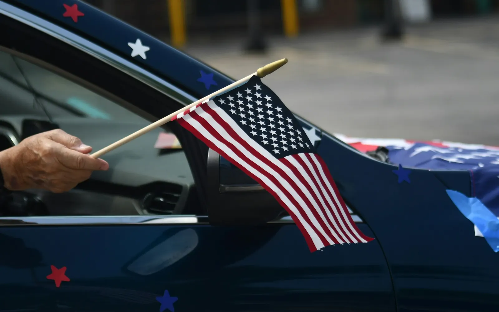 A hand holding a small American flag out of the window of a navy car decorated with star stickers.