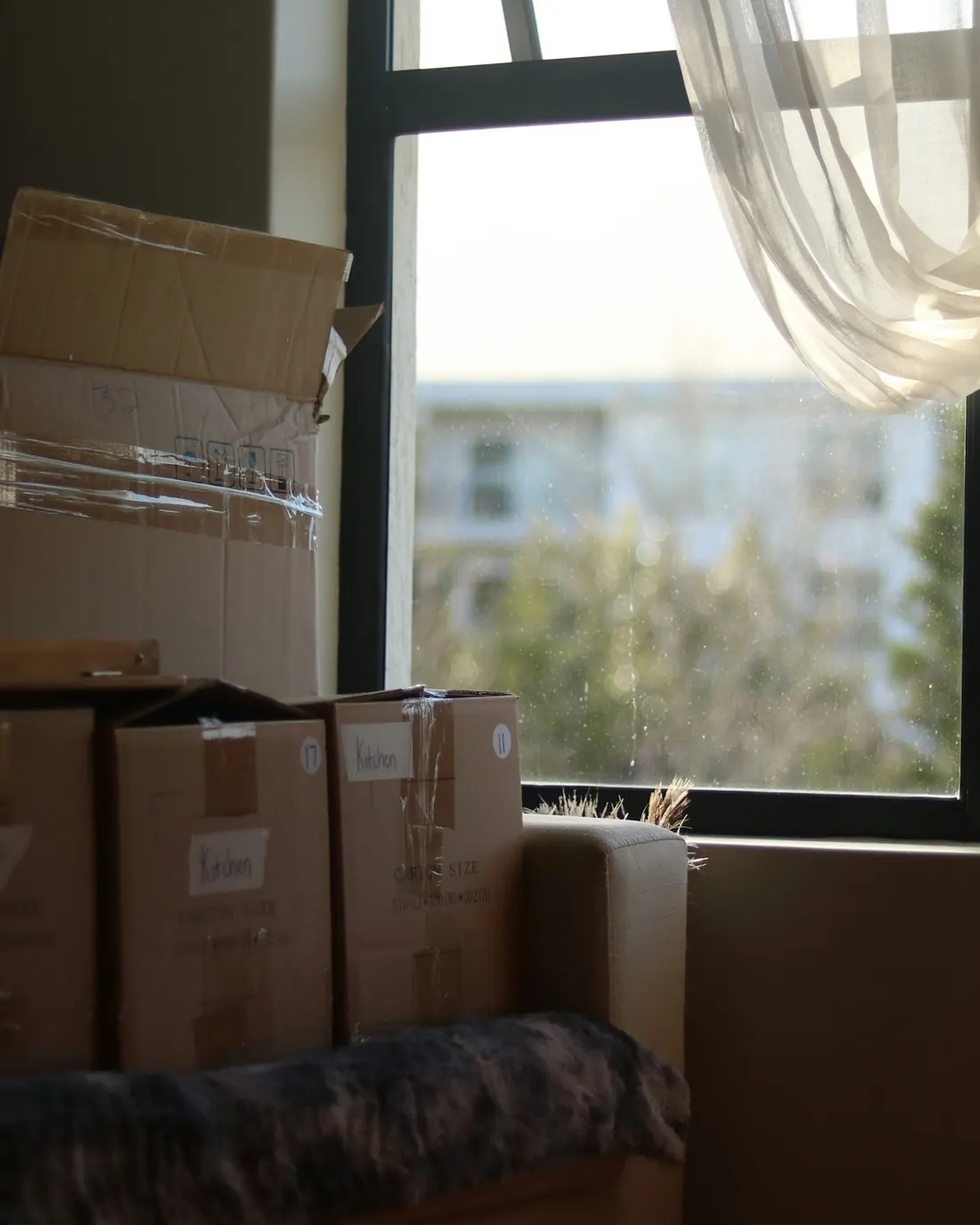Packed cardboard boxes labelled by room, stacked beside a window in an otherwise bare room.
