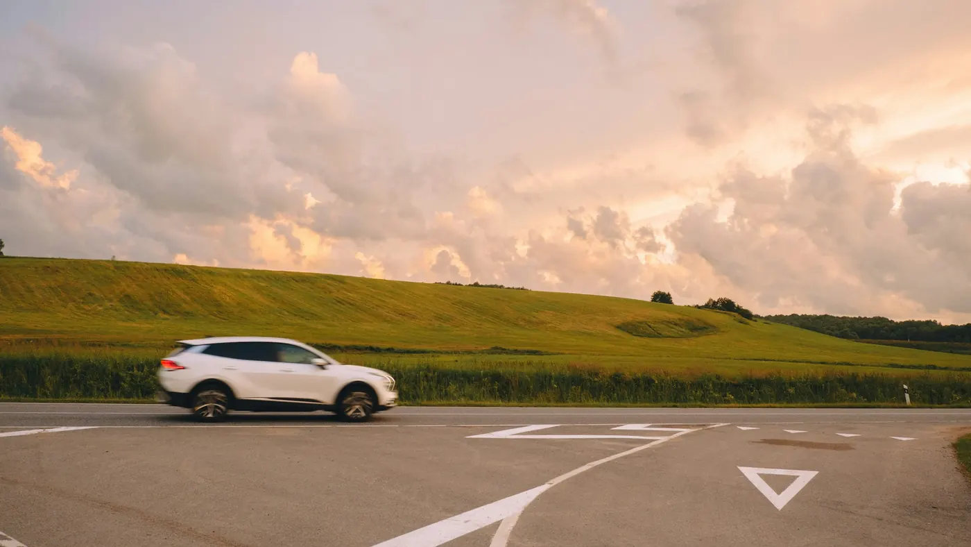 A white crossover crossing an empty road junction, with a green field and a tall bank of cloud behind it.