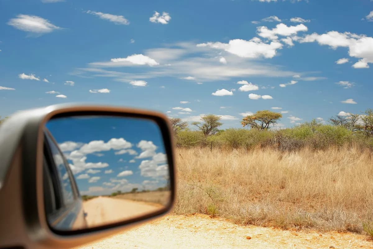 A car wing mirror reflecting a cloudy blue sky, beside dry grassland and low flat-topped trees.