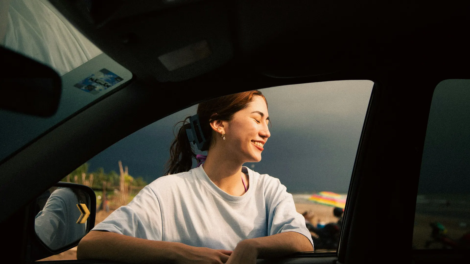 A passenger leaning out of an open car window into low sun, smiling.
